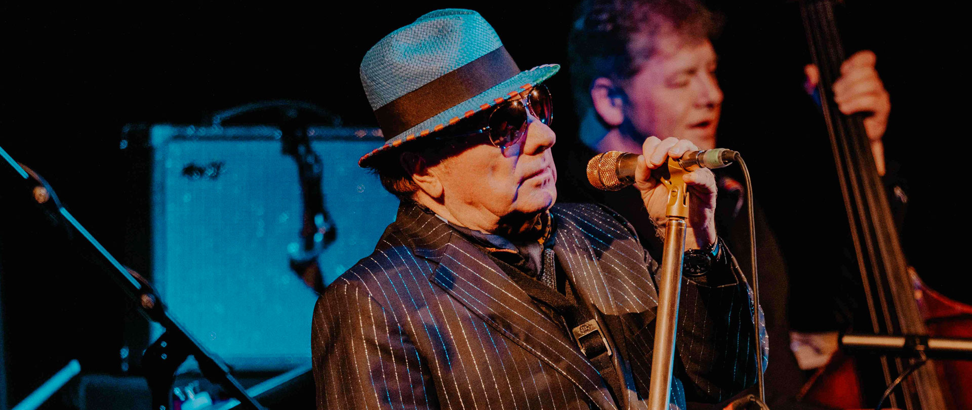 Van Morrison performing live, holding a microphone, wearing a blue hat and shades
